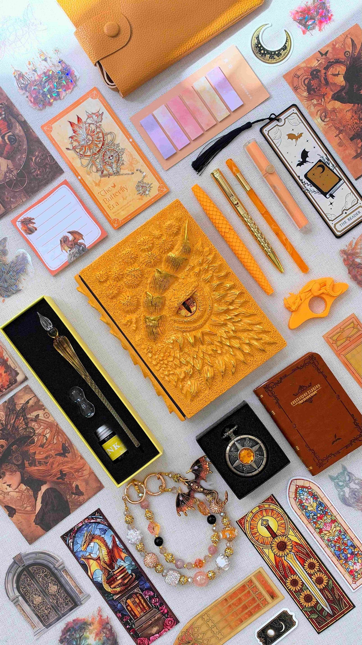 Amber Flame Linen Edition dragon bookish bundle in orange and gold, featuring a sculpted dragon eye notebook, dragon scale pen, glass dip pen and ink set, highlighter, small brown notebook, fantasy bookmarks, sticky notes, page tabs, dragon thumb page holder, beaded dragon charm, pocket watch and orange crossbody bag.