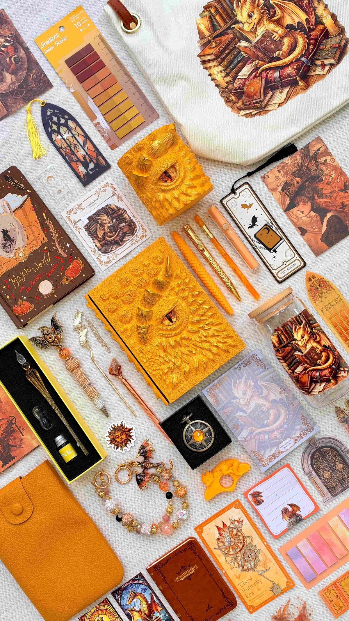 Amber Flame Velvet Edition dragon bookish bundle in orange and gold with a sculpted dragon eye notebook and matching pen holder organizer. Pens, a glass dip pen and ink set, fantasy bookmarks, sticky notes, reading tabs, dragon charm, pocket watch and notebooks surround a dragon print glass jar, dragon and books tote bag and orange crossbody bag.
