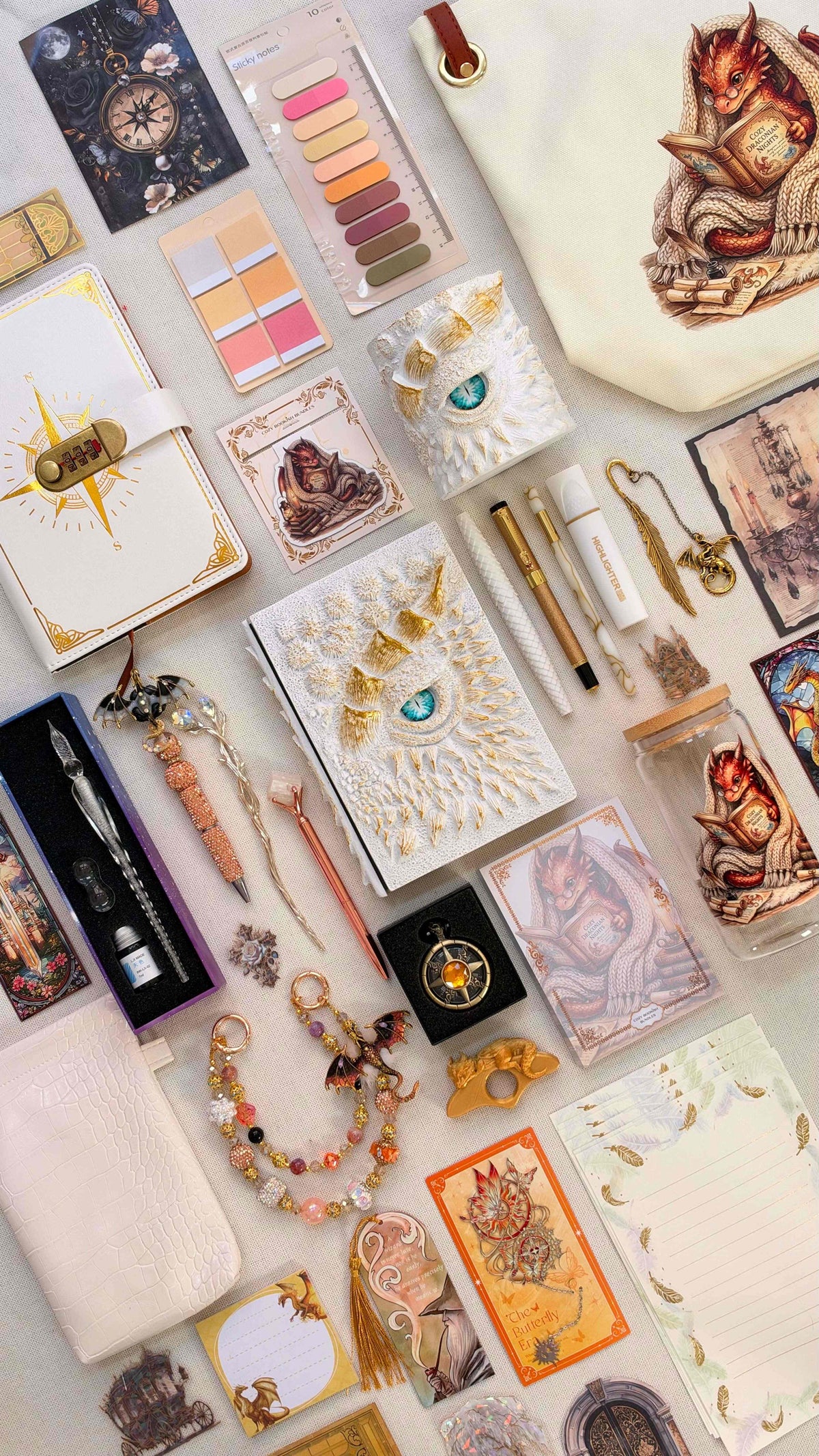 White & Gold Velvet Edition dragon bookish bundle with a white and gold dragon eye notebook and matching pen holder organizer. White and gold pens, a glass dip pen and ink set, fantasy bookmarks, writing paper, sticky notes, reading tabs, pocket watch and dragon charm sit beside an extra notebook, white crossbody bag, dragon print glass jar and dragon tote bag.