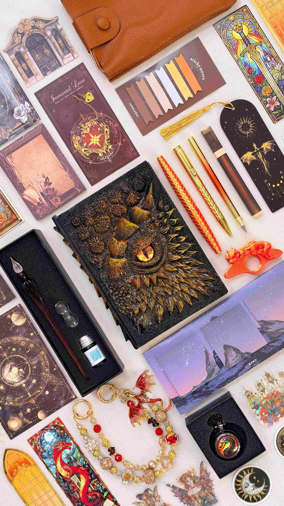Gilded Ember Linen Edition dragon bookish bundle with a black and gold sculpted dragon eye notebook, orange and gold pens, glass dip pen and ink set, fantasy bookmarks, sticky notes, page tabs, dragon thumb page holder, beaded dragon charm, pocket watch and orange crossbody bag.
