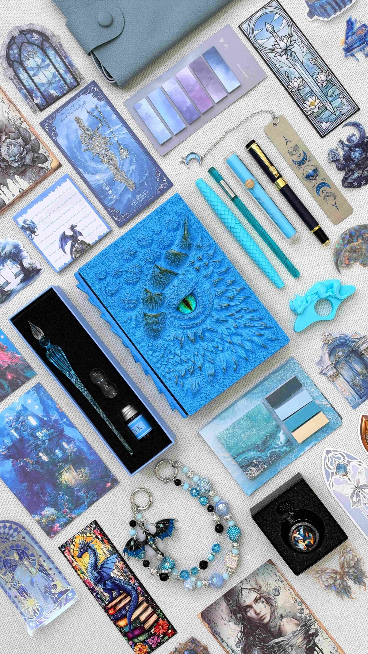 Ice Crystal Linen Edition blue dragon bookish bundle: a sculpted dragon eye notebook, dragon scale pen, glass dip pen with ink, highlighter, fantasy bookmarks, sticky notes, page tabs, dragon thumb page holder, beaded dragon charm, pocket watch and pale blue crossbody bag.