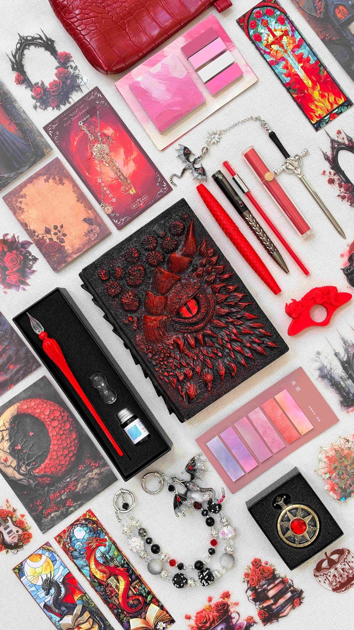 Inferno Eye Linen Edition black and red dragon bookish bundle with a sculpted dragon eye notebook, red dragon scale pen, glass dip pen and ink set, highlighter, fantasy bookmarks, sticky notes, pink page tabs, dragon thumb page holder, beaded dragon charm, pocket watch and red crossbody bag.