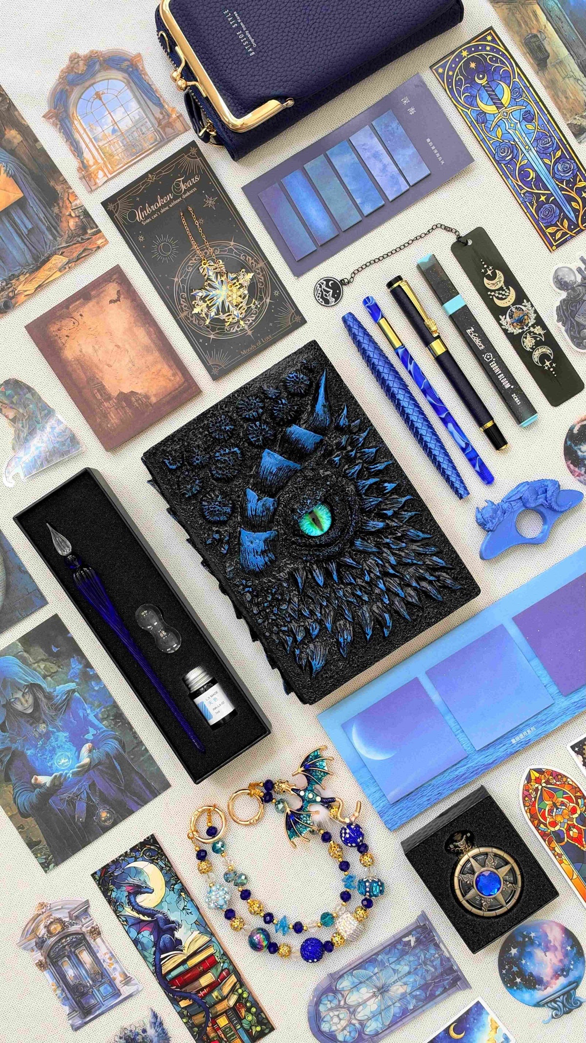 Midnight Linen Edition dragon bookish bundle in black and blue, featuring a sculpted dragon eye notebook with a green eye, blue pens, glass dip pen and ink set, highlighter, fantasy bookmarks, sticky notes, page tabs, dragon thumb page holder, beaded dragon charm, pocket watch and navy crossbody bag.