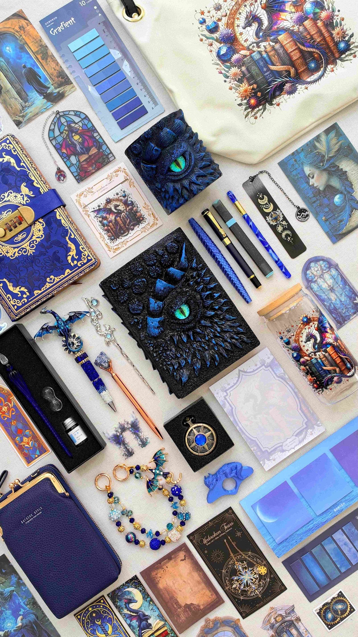 Midnight Velvet Edition black and blue dragon bookish bundle with a sculpted dragon eye notebook and matching pen holder organizer. Pens, a glass dip pen set, fantasy bookmarks, sticky notes and reading tabs sit beside an extra notebook, dragon print tote bag and glass jar, navy crossbody bag, dragon charm and pocket watch.