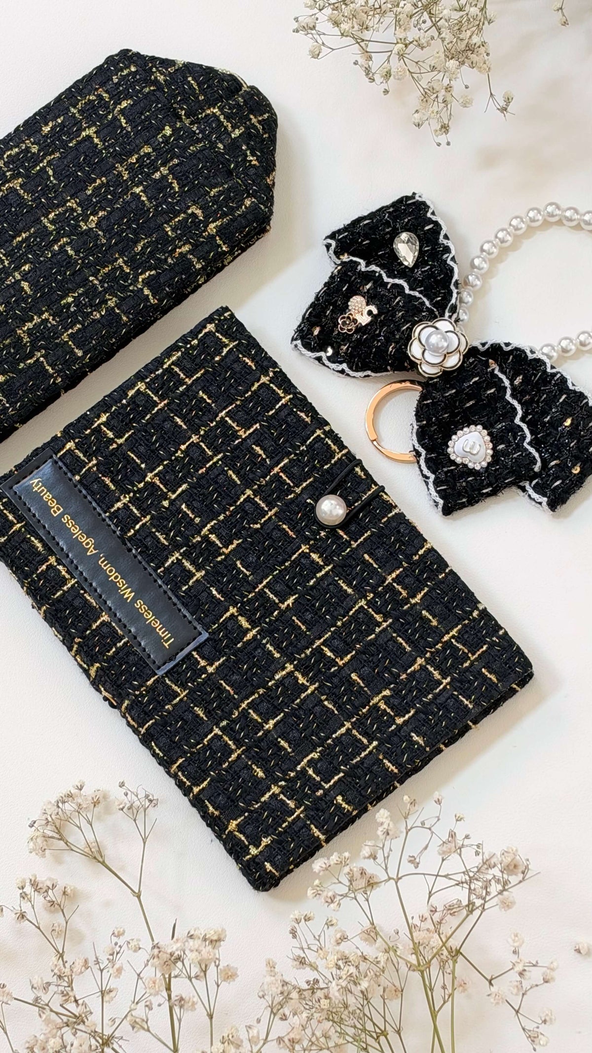 Close-up of the Noir Linen Edition: a black-and-gold tweed notebook and pouch with a pearl-detailed bow on a pale floral surface.