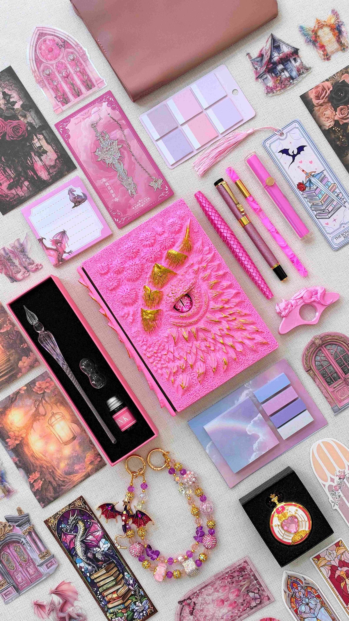 Pink Fantasy World Linen Edition dragon bookish bundle with a bright pink and gold sculpted dragon eye notebook, pink pens and highlighter, glass dip pen and ink set, fantasy bookmarks, sticky notes, pastel page tabs, dragon thumb page holder, beaded dragon charm, pocket watch and dusty pink crossbody bag.
