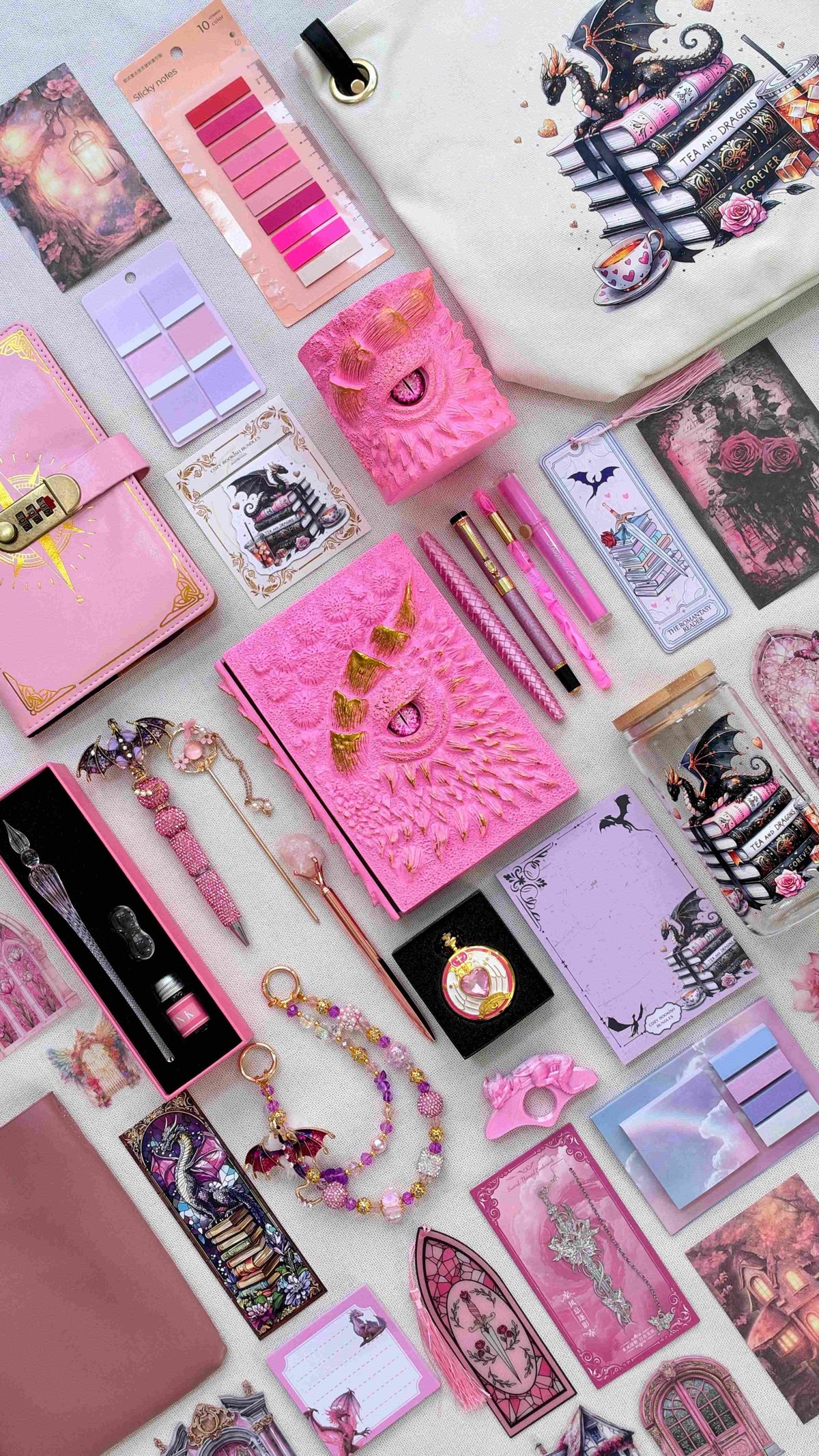 Pink Fantasy World Velvet Edition dragon bookish bundle with a pink and gold dragon eye notebook and matching pen holder organizer. Flat lay includes pink pens, a glass dip pen and ink set, fantasy bookmarks, sticky notes, reading tabs, dragon charm, pocket watch, extra notebook, crossbody bag, dragon print glass jar and dragon and books tote bag.