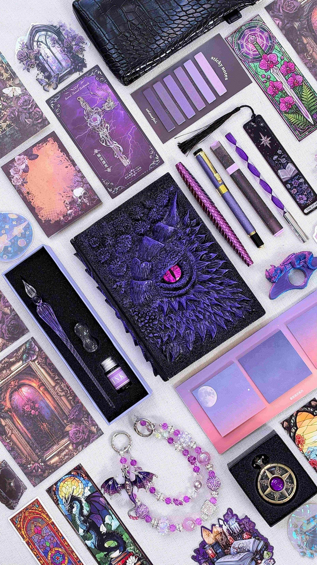 Purple Shadow Eye Linen Edition black and purple dragon bookish bundle with a sculpted dragon eye notebook and pink eye, purple pens, glass dip pen and ink set, fantasy bookmarks, sticky notes, page tabs, dragon thumb page holder, beaded dragon charm, pocket watch and black crossbody bag.