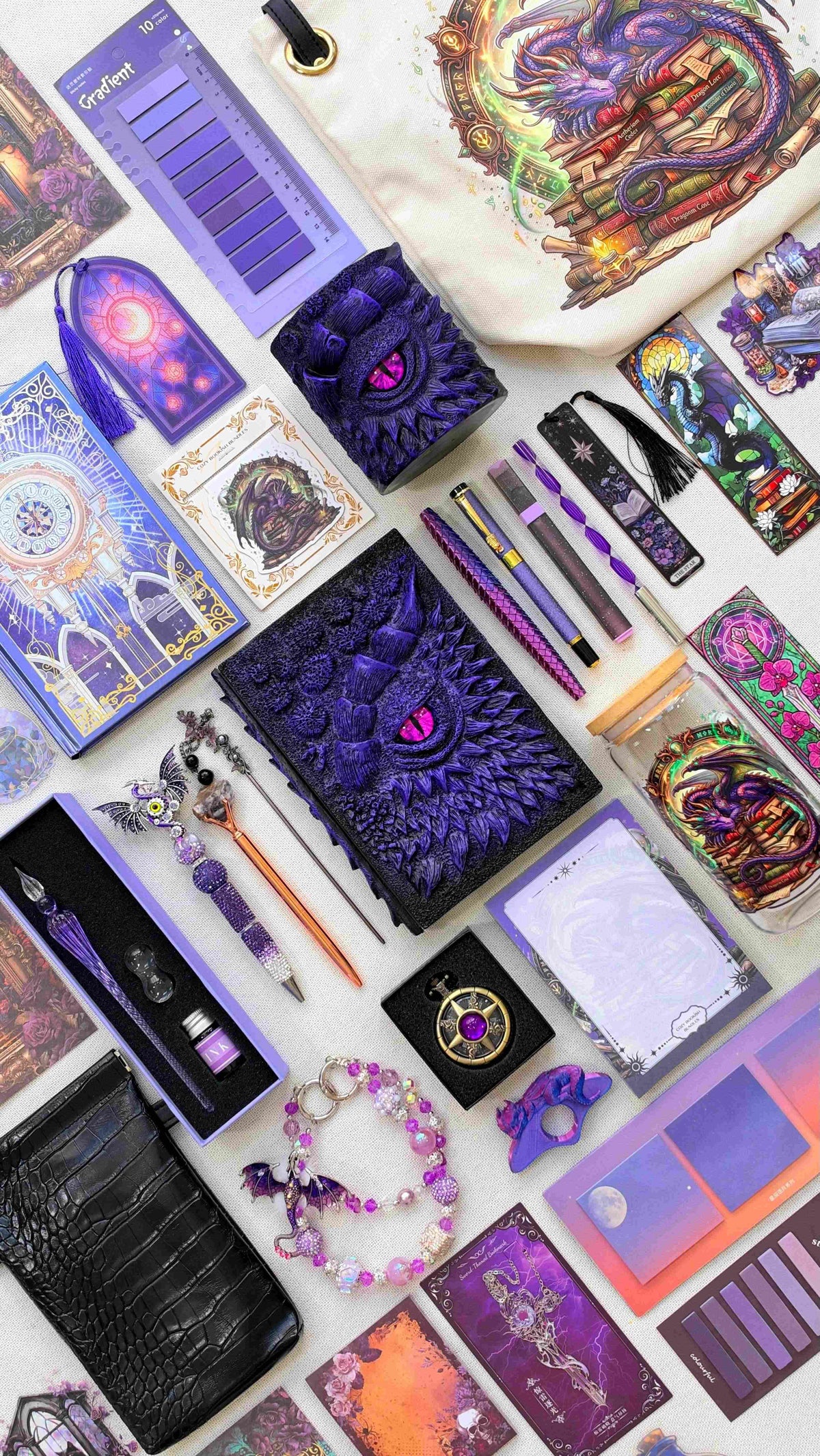 Purple Shadow Eye Velvet Edition black and purple dragon bookish bundle with a sculpted dragon eye notebook and matching pen holder organizer. Purple pens, a glass dip pen set, fantasy bookmarks, sticky notes and reading tabs surround an extra notebook, dragon print tote bag and glass jar, black crossbody bag, dragon charm and pocket watch.