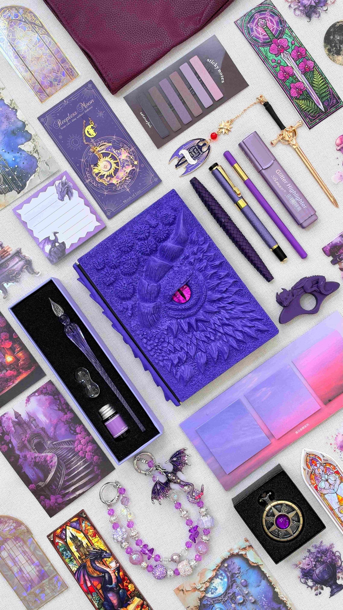 Royal Amethyst Linen Edition dragon bookish bundle in deep purple, arranged with a sculpted dragon eye notebook, dragon scale pen, glass dip pen and ink set, highlighter, fantasy bookmarks, sticky notes, page tabs, dragon thumb page holder, beaded dragon charm, pocket watch and burgundy crossbody bag.