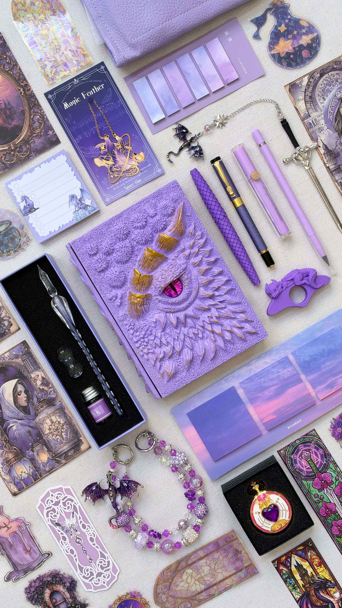 Soft Purple Linen Edition dragon bookish bundle with a lilac sculpted dragon eye notebook, gold horns and a pink eye. Arranged with purple pens and highlighter, a glass dip pen and ink set, fantasy bookmarks, sticky notes, pastel page tabs, dragon thumb page holder, beaded dragon charm, pocket watch and lavender crossbody bag.