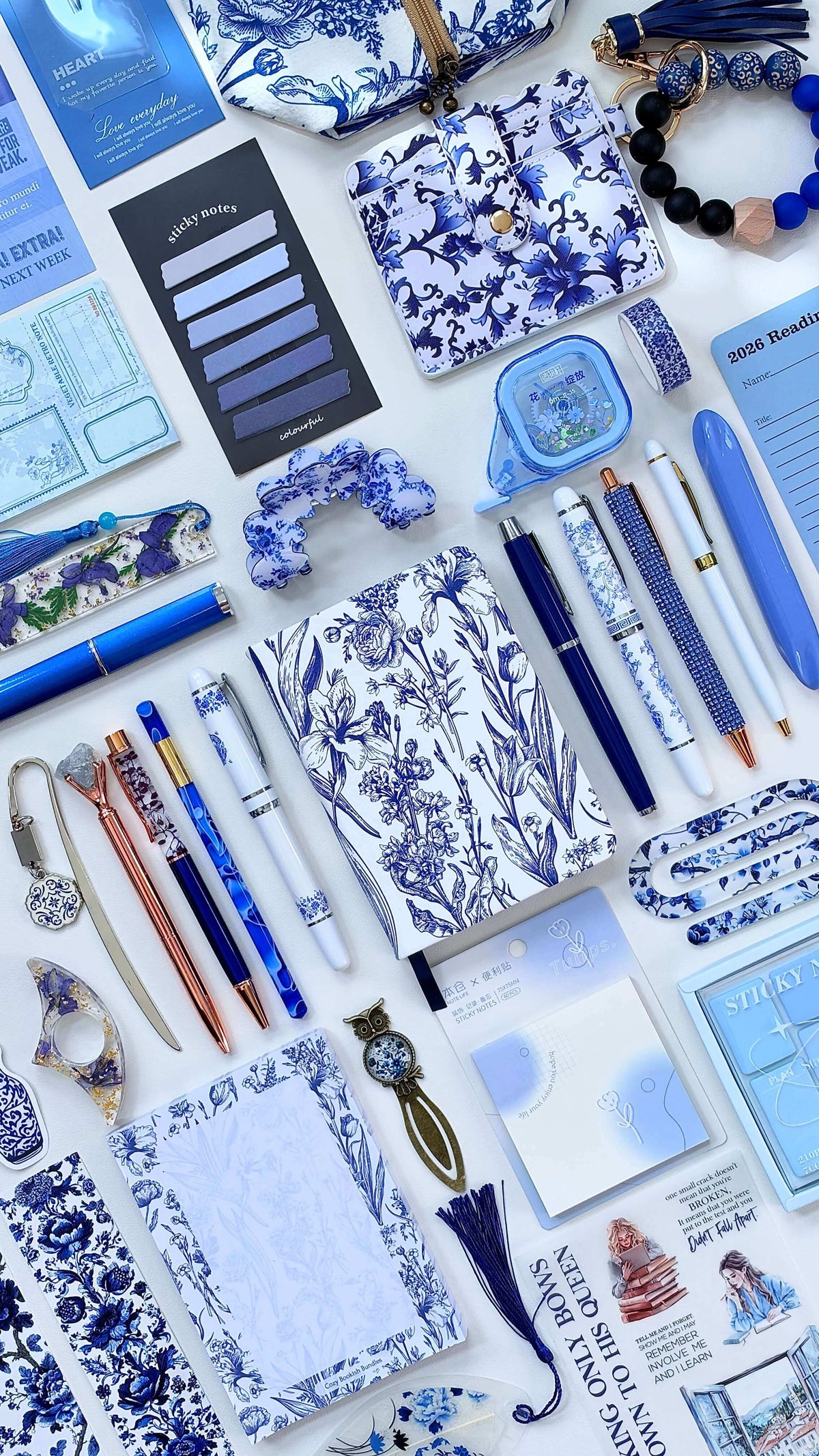 A flat-lay arrangement of blue and white themed stationery and accessories, including patterned notebooks, pens, sticky notes, bracelets, and small decorative items on a white background. Blue and white Toile de Jouy bookish bundle with a floral journal, pens, bookmarks, sticky notes