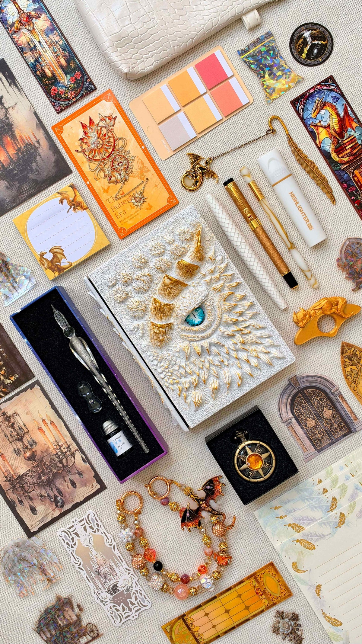 White & Gold Linen Edition dragon bookish bundle with a white sculpted dragon eye notebook, gold horns and a blue eye. Flat lay includes white and gold pens, a glass dip pen and ink set, highlighter, fantasy bookmarks, decorative letter writing paper, sticky notes, page tabs, dragon thumb page holder, beaded dragon charm, pocket watch and white crossbody bag.